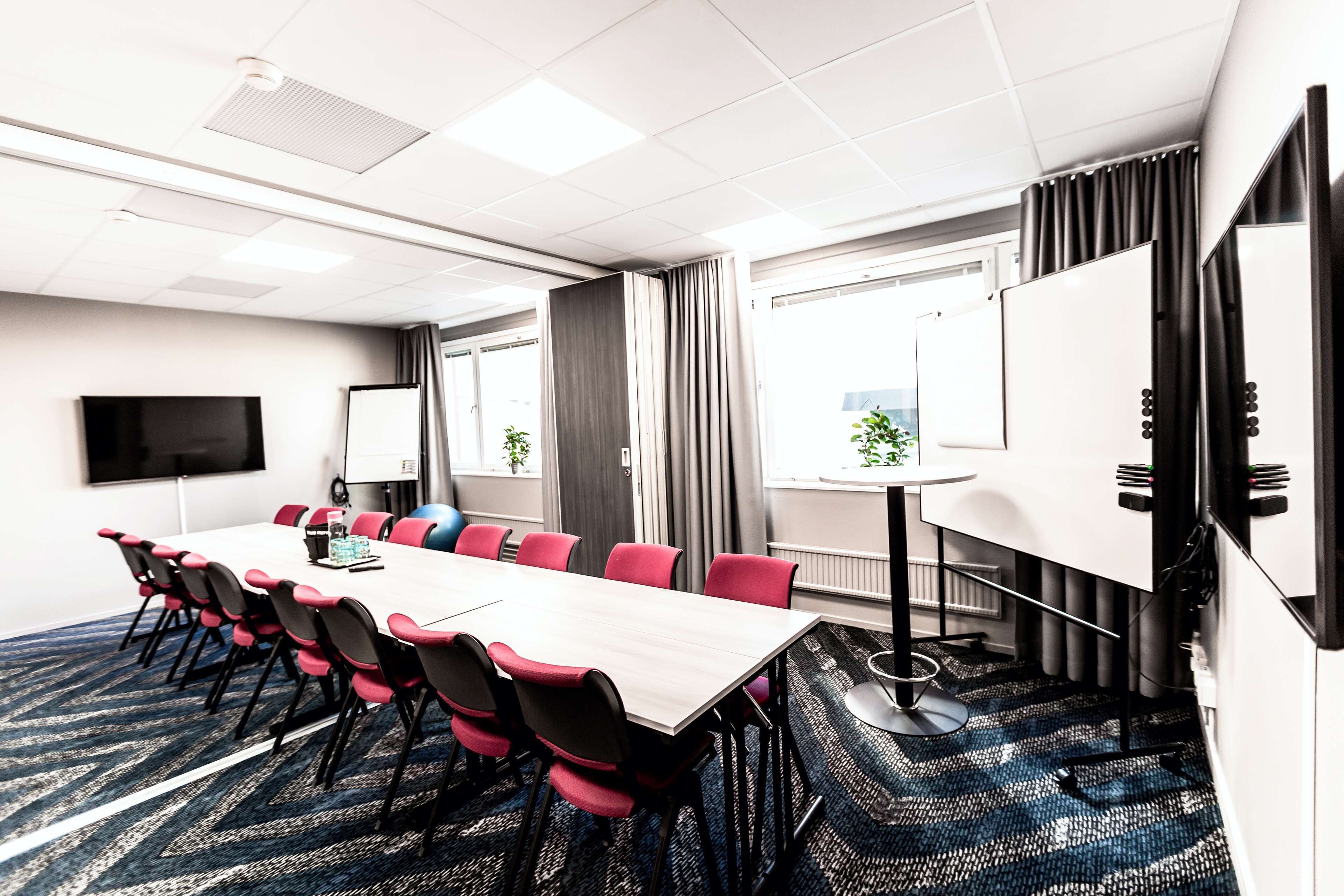 A conference room with a long table, red and black chairs, a whiteboard, and a projector screen.