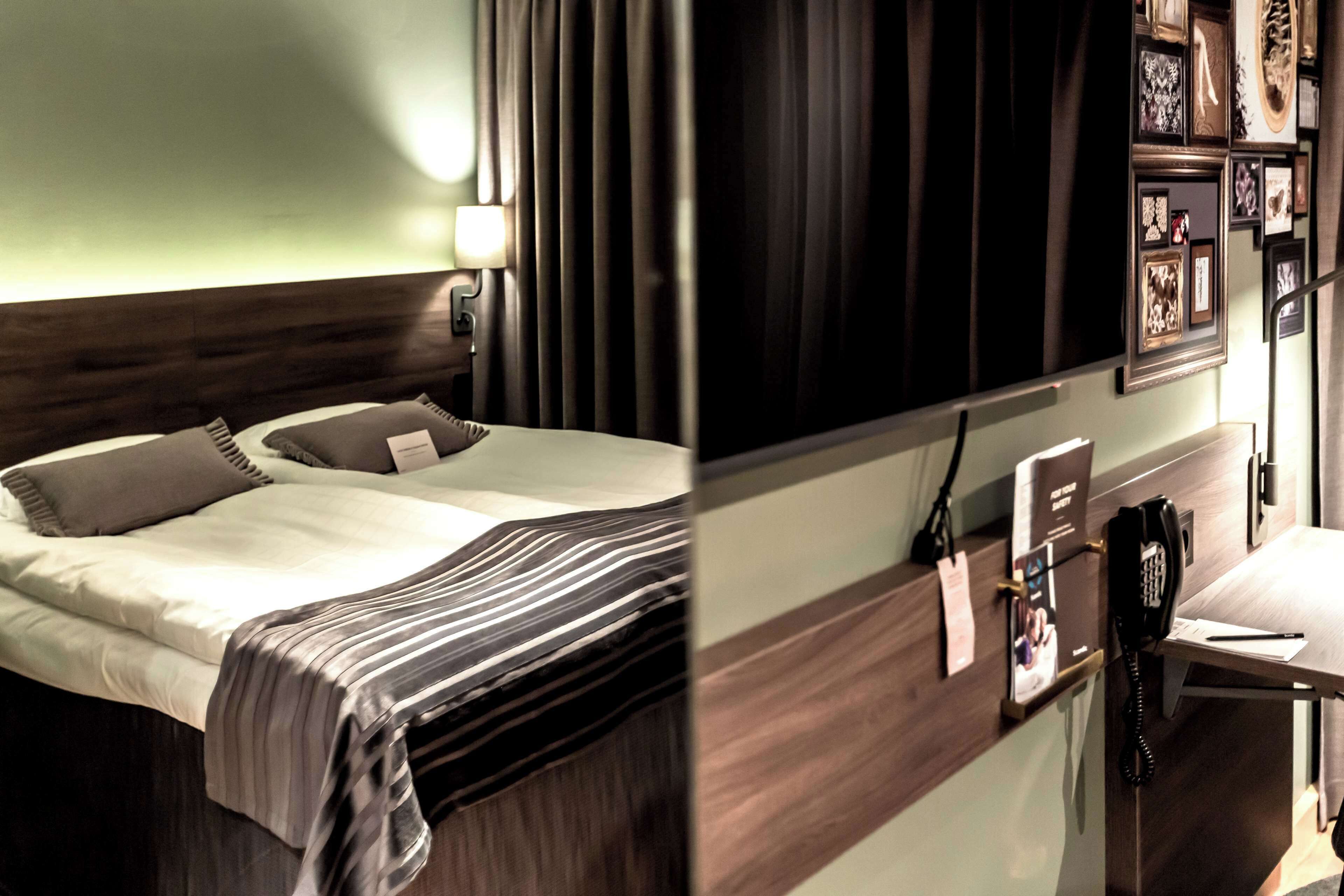 Ett hotellrum med en säng, ett sängbord, en lampa och inramade tavlor på väggen.