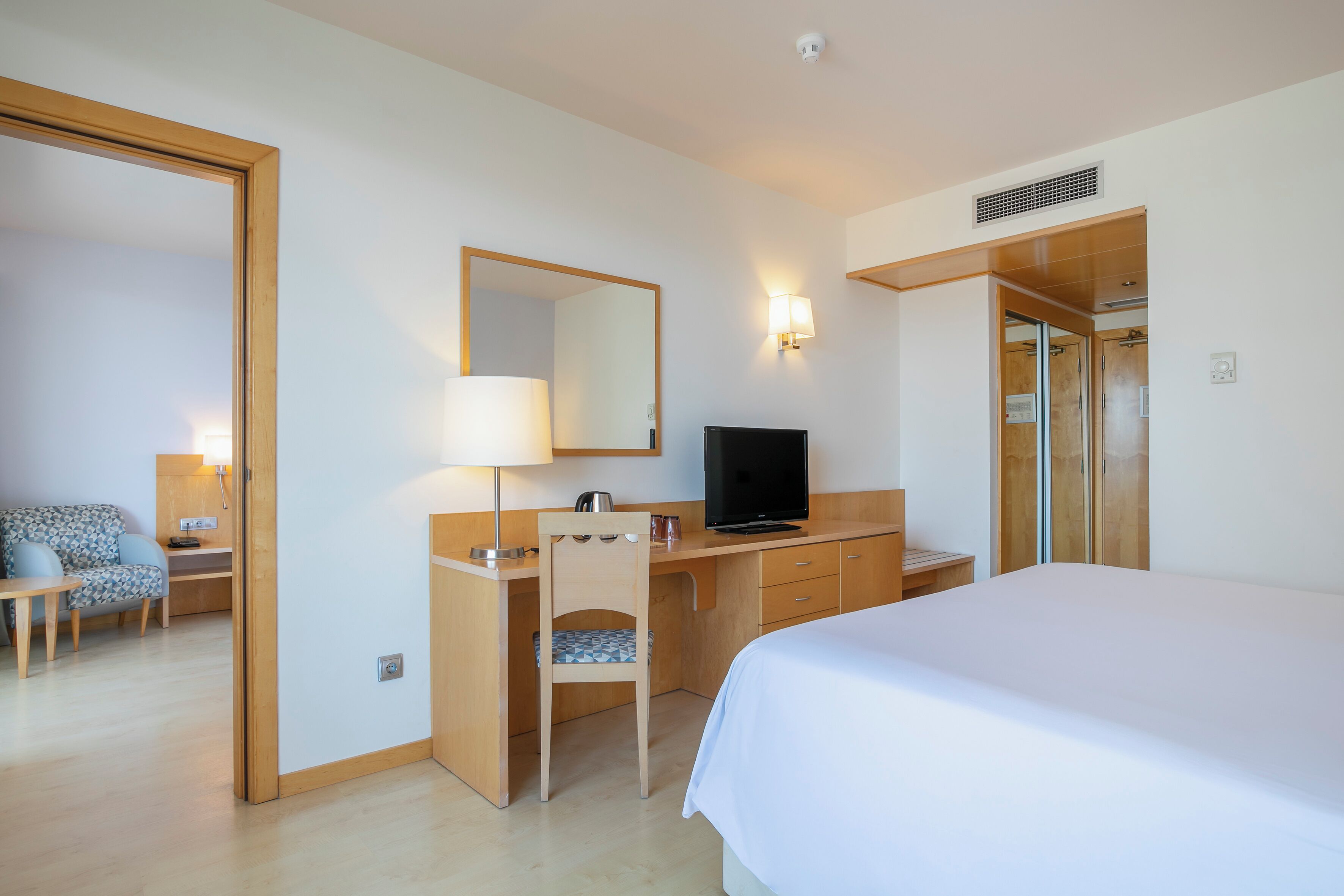 A hotel room with a bed, a desk with a chair, a television, a mirror, and a wardrobe.
