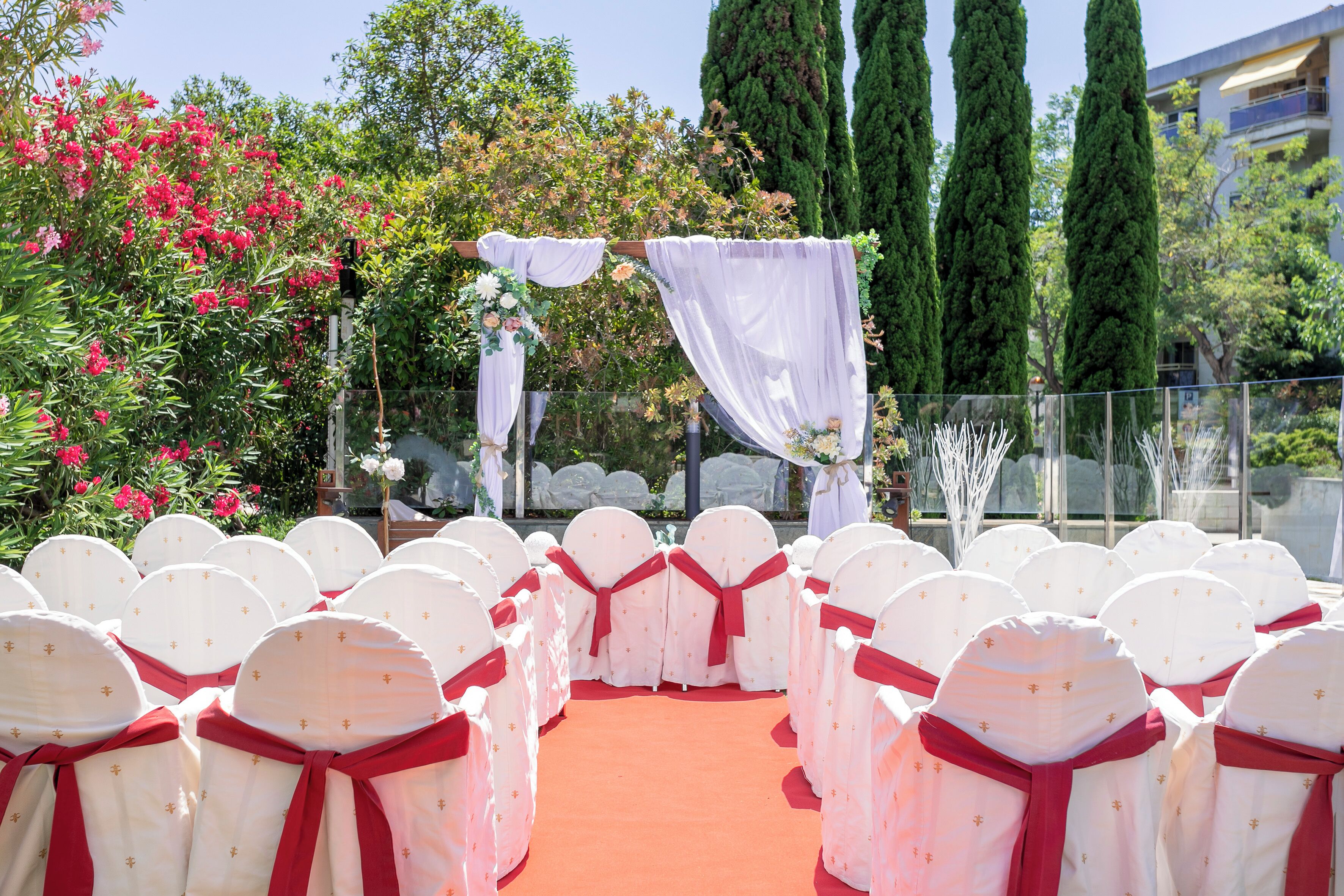 Un pasillo con alfombra roja, sillas blancas y lazos rojos, que conduce a un espacio al aire libre para una ceremonia, con cortinas blancas y arreglos florales.