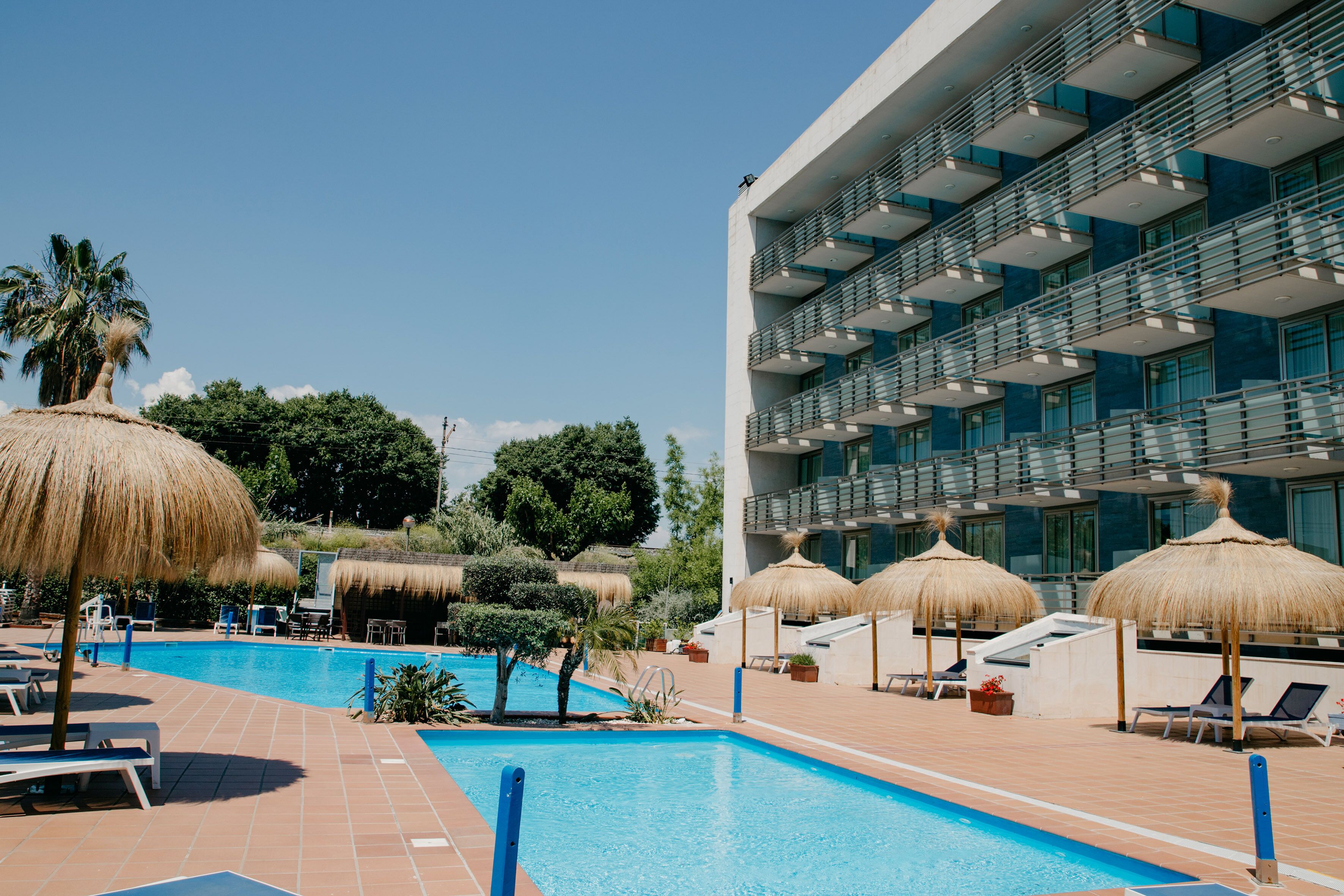 A swimming pool area with lounge chairs and thatched umbrellas, surrounded by a modern building with balconies.