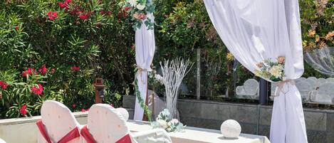 An outdoor wedding setup with white chairs, a table, and a backdrop of greenery and flowers.