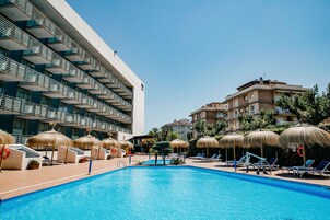 A swimming pool with lounge chairs and thatched umbrellas, surrounded by buildings with balconies and a clear sky.