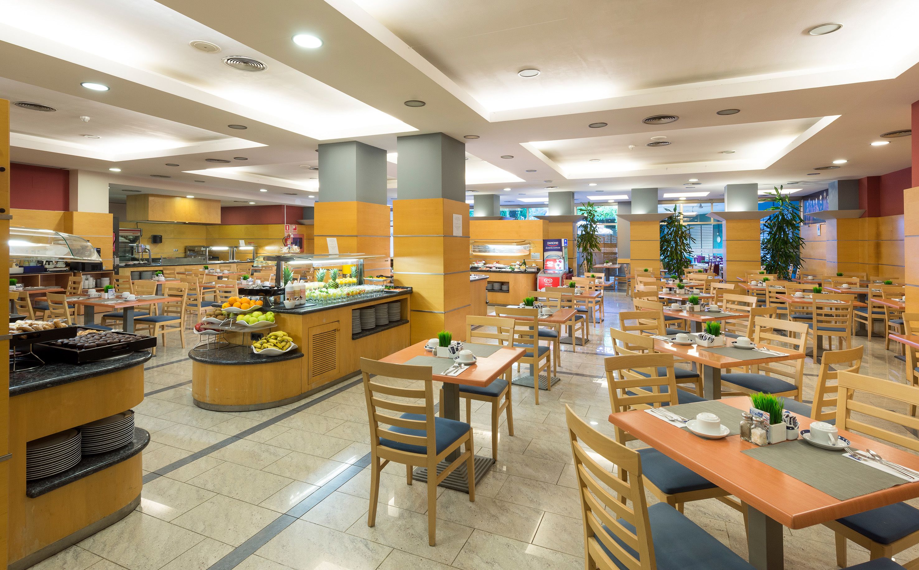 A spacious cafeteria with multiple tables and chairs, a central buffet area with various food items, and a bright, well-lit environment.