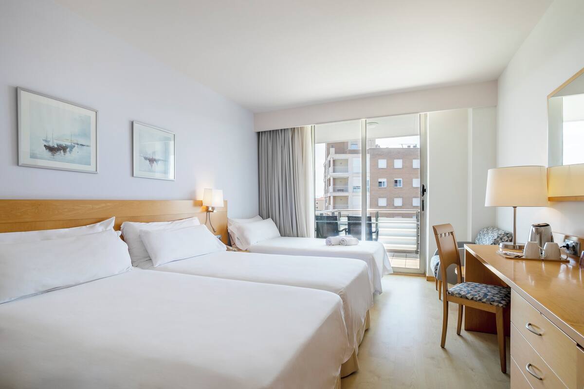 Une chambre d’hôtel avec deux lits, un bureau et une chaise. Il y a deux tableaux encadrés au mur, une fenêtre avec des rideaux et une vue sur les bâtiments à l’extérieur.
