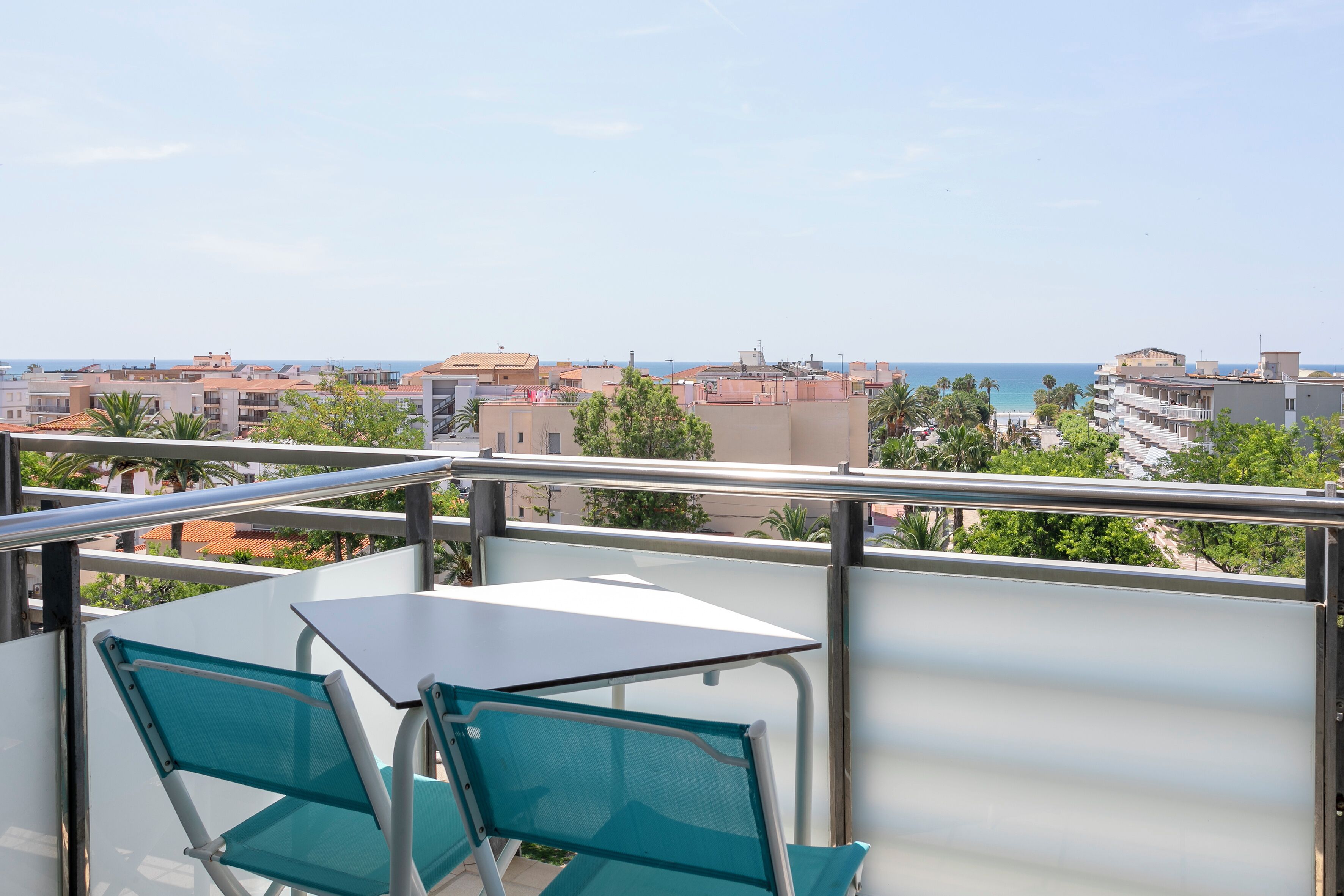 A balcony with a table and chairs, overlooking a coastal cityscape with buildings and a beach.
