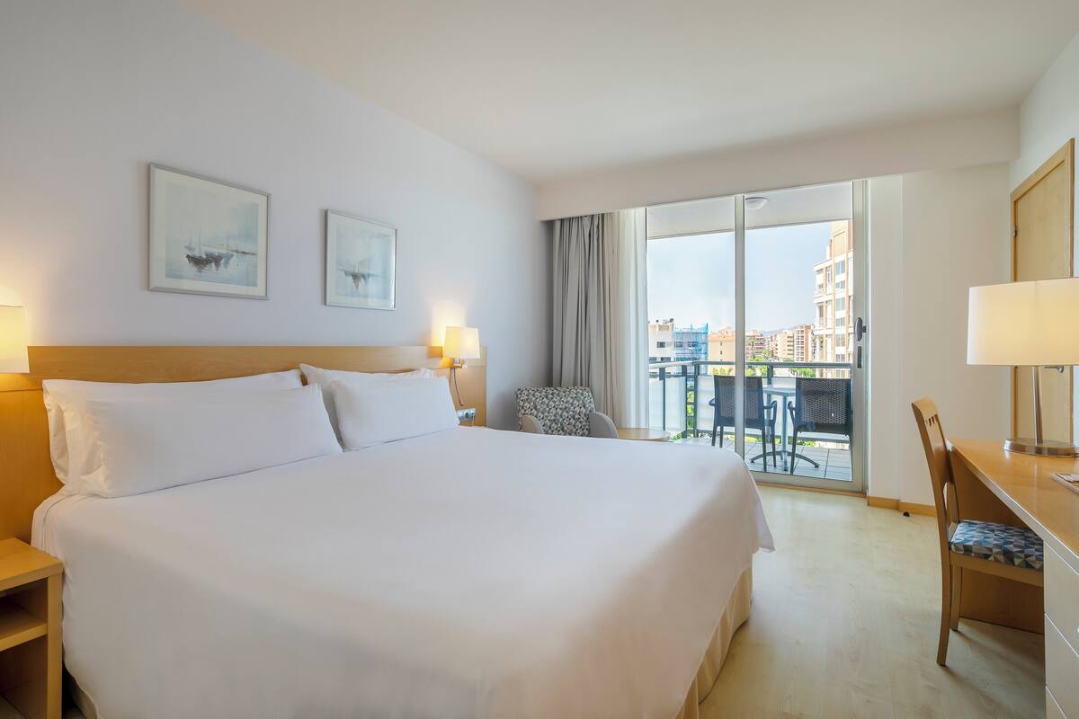 Une chambre d’hôtel avec un grand lit, deux tables de chevet avec des lampes, une chaise, un bureau avec une chaise et un balcon donnant sur des immeubles.
