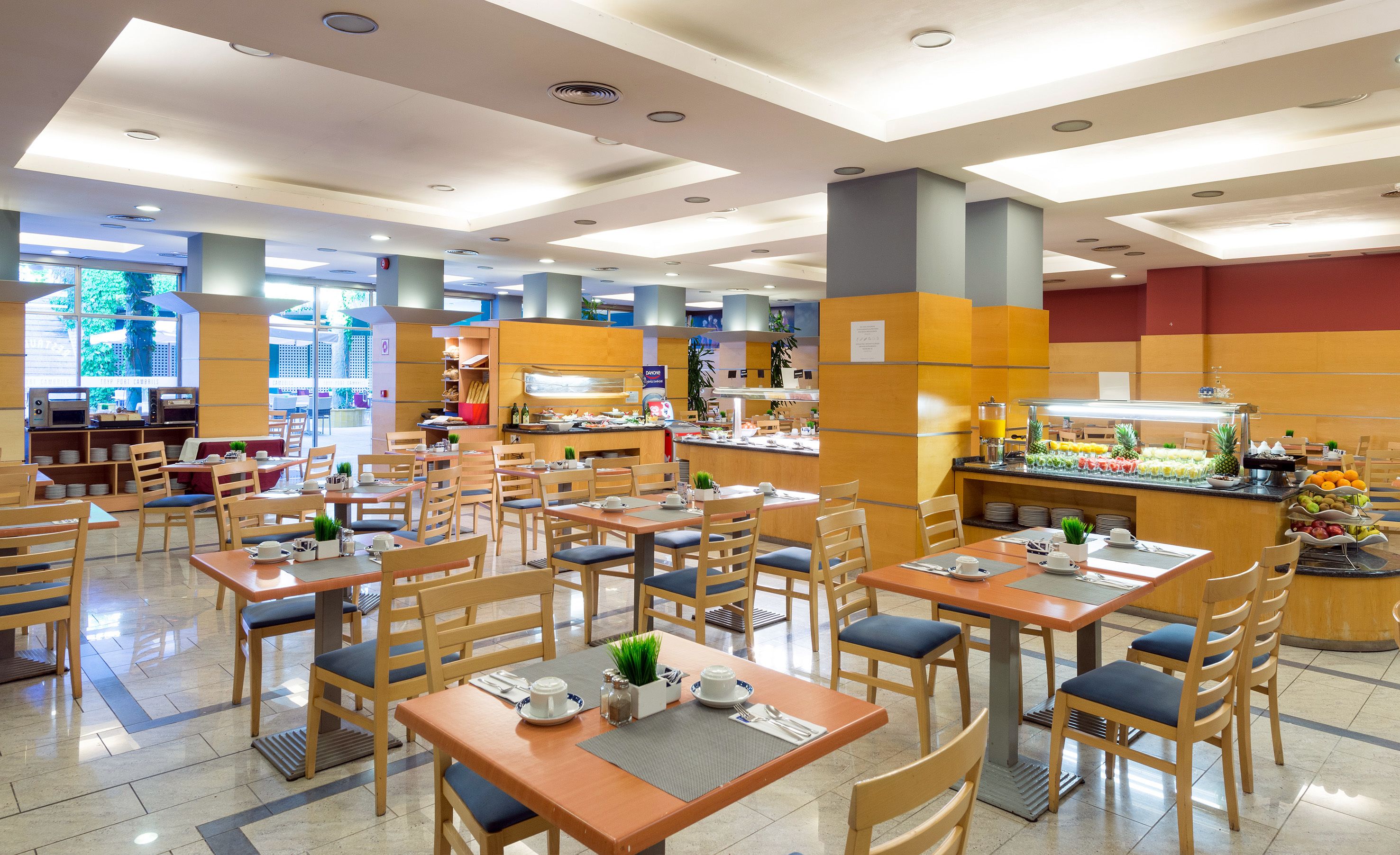 A spacious dining area with multiple tables and chairs, a buffet counter with various food items, and a view of the outside through large windows.