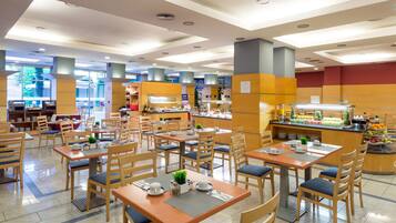 A spacious dining area with multiple tables and chairs, a buffet counter with various food items, and a view of the outside through large windows.