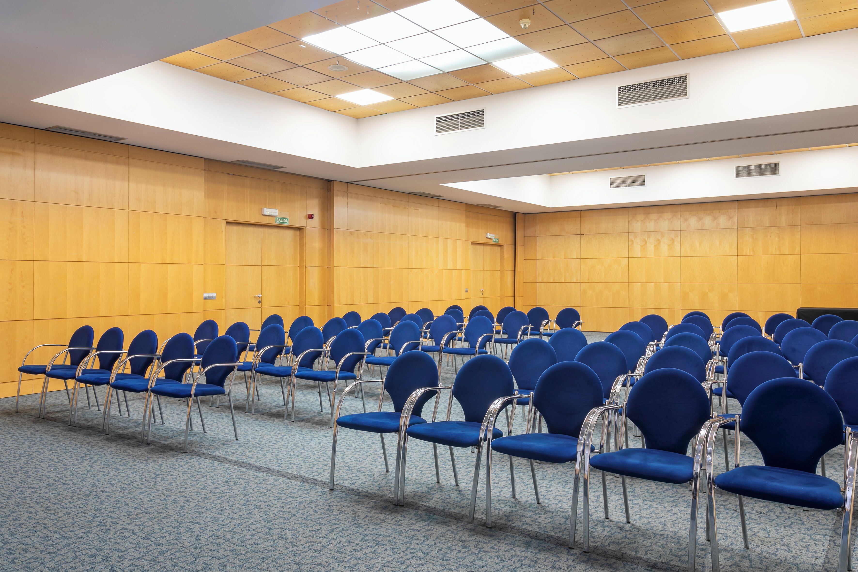 A conference room with rows of blue chairs and a wooden panel wall.