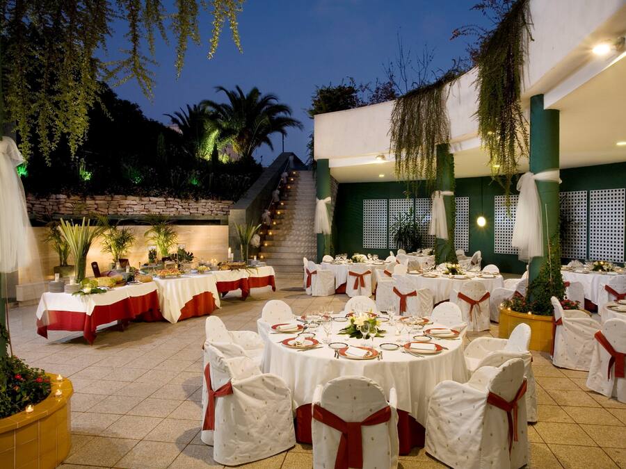Un salón de banquetes con mesas redondas, manteles blancos y cintas rojas, rodeado de follaje y palmeras.