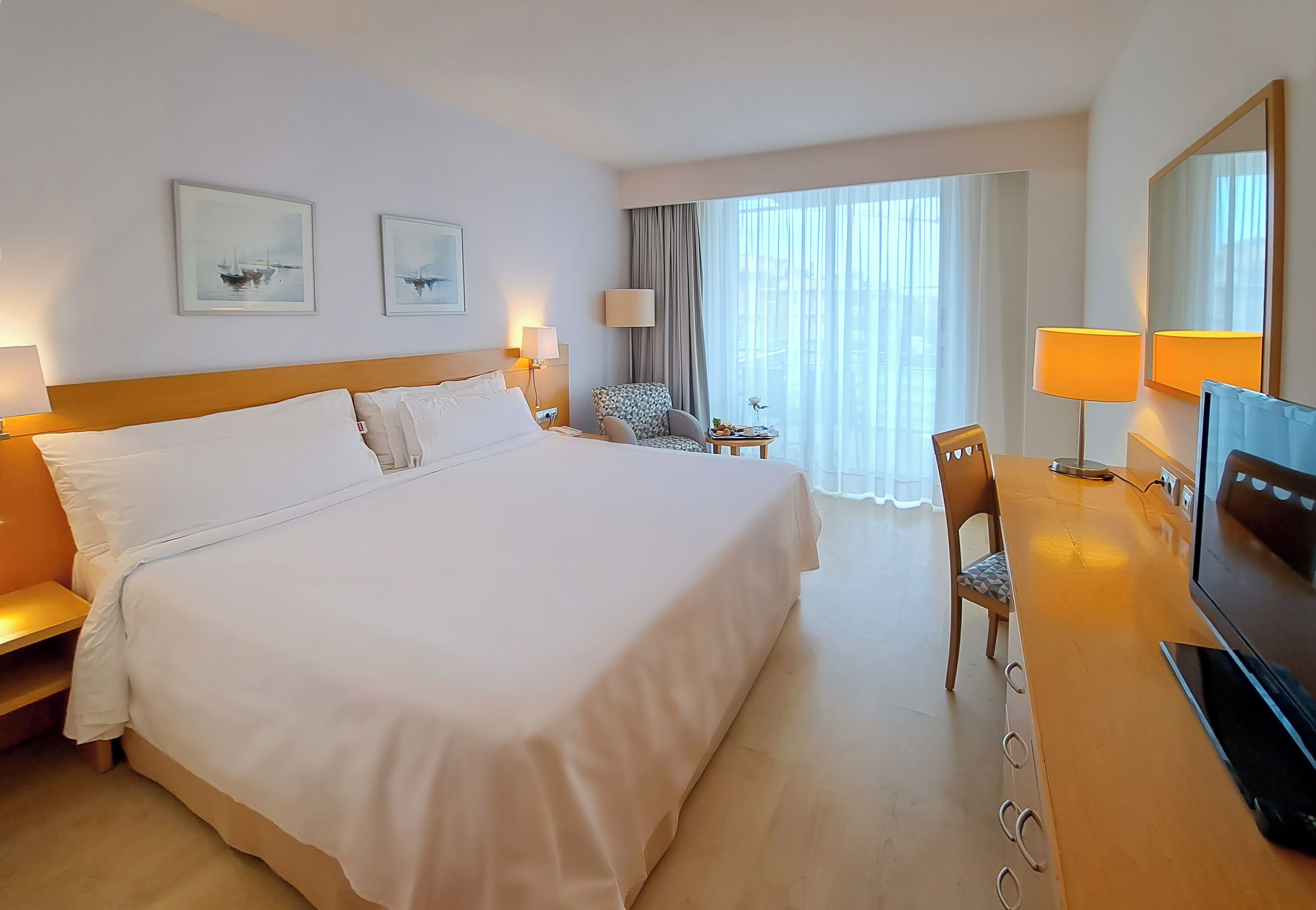 Une chambre d’hôtel avec un grand lit, un bureau avec une télévision, deux chaises, une lampe et une fenêtre avec des rideaux.