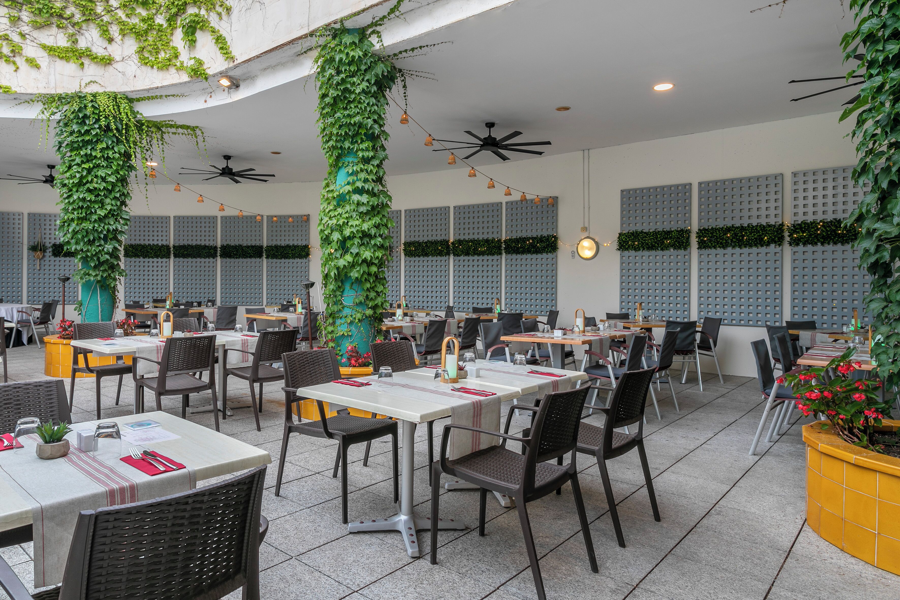 Un espace extérieur aménagé pour les repas, avec des tables et des chaises, des ventilateurs de plafond et des plantes décoratives.