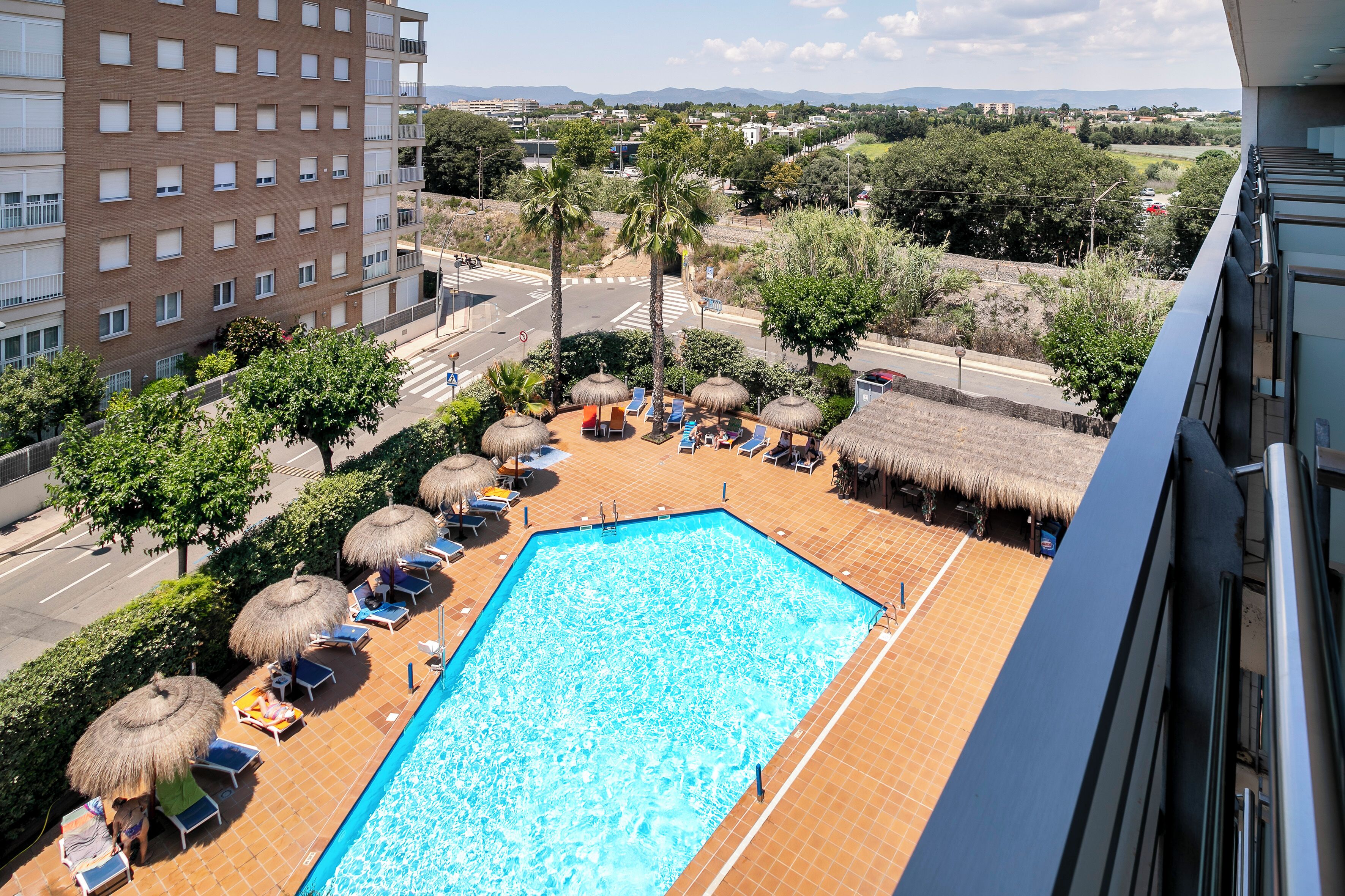Una piscina en la azotea con sillones de descanso y sombrillas, rodeada de vegetación y vistas a edificios lejanos.