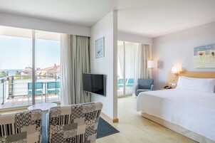 A hotel room with a bed, a TV, a chair, a table, and a balcony with a view of the sea.
