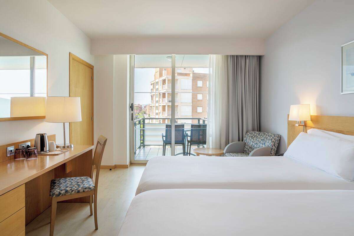 Une chambre d’hôtel équipée d’un lit, d’un bureau, d’une chaise et d’un balcon donnant sur des immeubles.