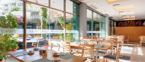 Een ruim café met houten tafels en stoelen, grote ramen met planten en uitzicht op de straat.