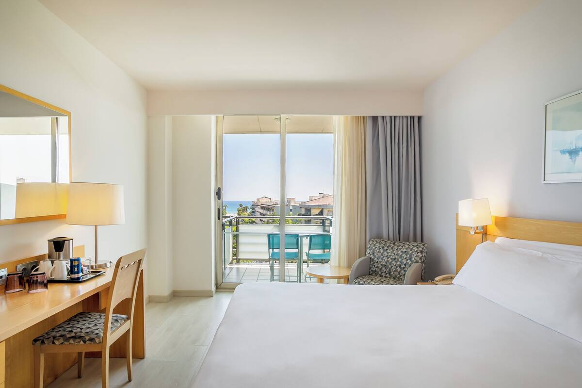 Une chambre d’hôtel avec un lit, un bureau doté d’une lampe, une chaise, une petite table et une vue sur la mer, accessible par une porte coulissante.