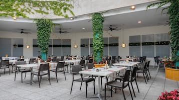 An outdoor dining area with tables and chairs, ceiling fans, and greenery.