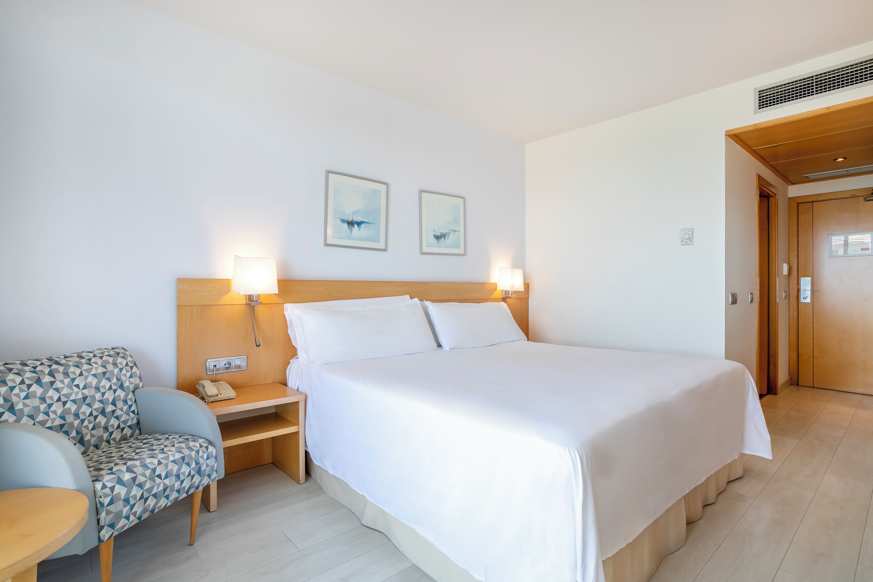 A hotel room with a bed, a bedside table with a lamp, a chair, and two wall-mounted artworks.
