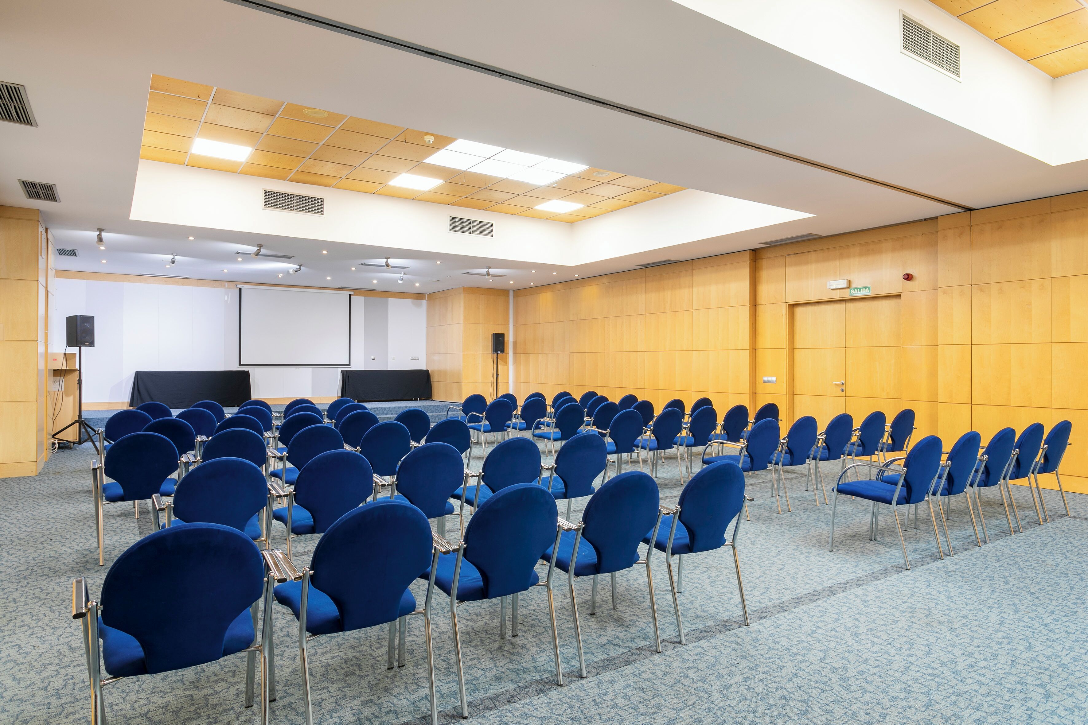 Un salón de conferencias con filas de sillas azules, una pantalla de proyector y un panel de pared de madera.