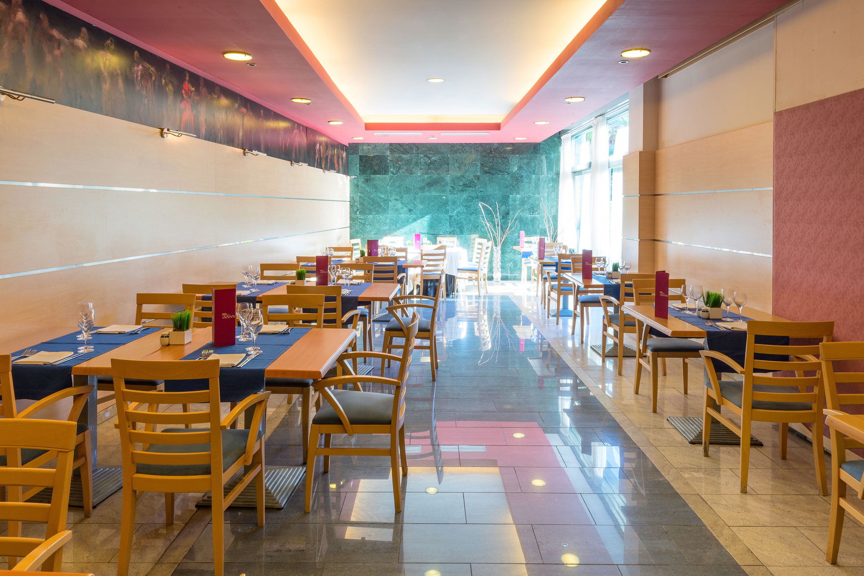 Een ruim restaurant met meerdere tafels en stoelen, een marmeren wand en uitzicht door de ramen.