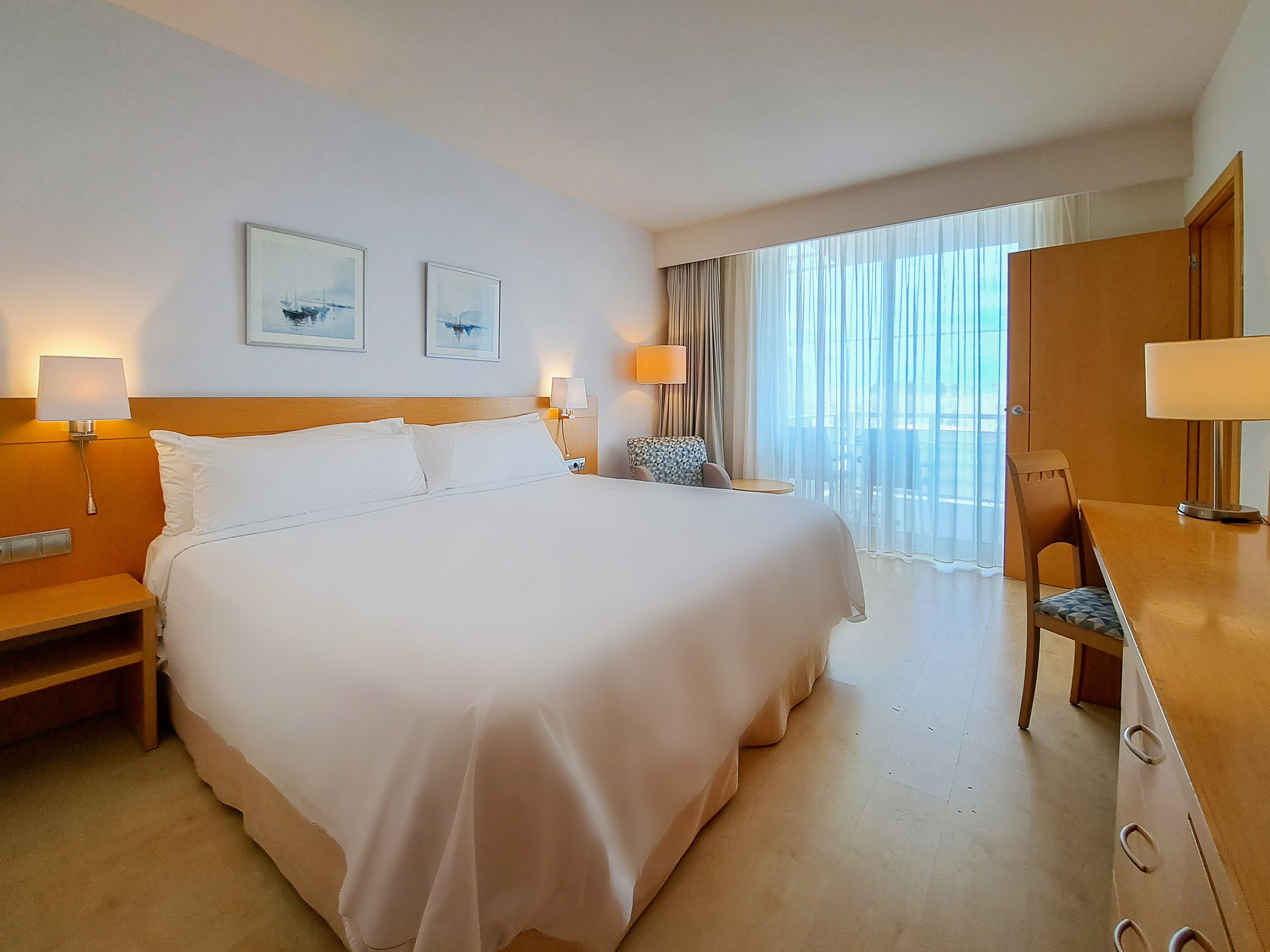 Ein Hotelzimmer mit einem großen Bett, Nachttischen inklusive Lampen, einem Schreibtisch mit Stuhl und einem großen Fenster mit Vorhängen.