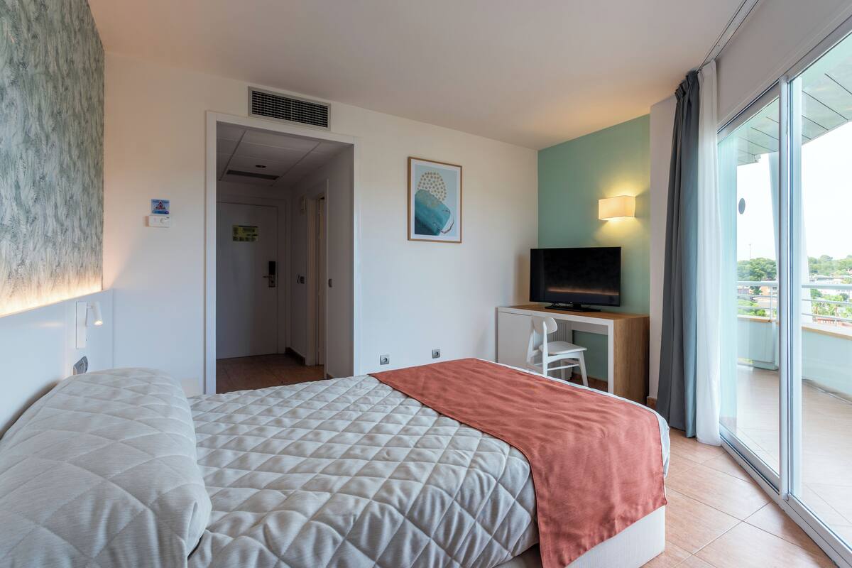 A hotel room with a bed, a desk with a TV, a window with a view, and a door leading to another room.