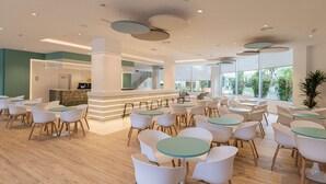 A spacious indoor area with multiple round tables and white chairs, a reception desk, and large windows offering a view of greenery outside. - Ponient Pirámide Salou by PortAventura World (Salou)