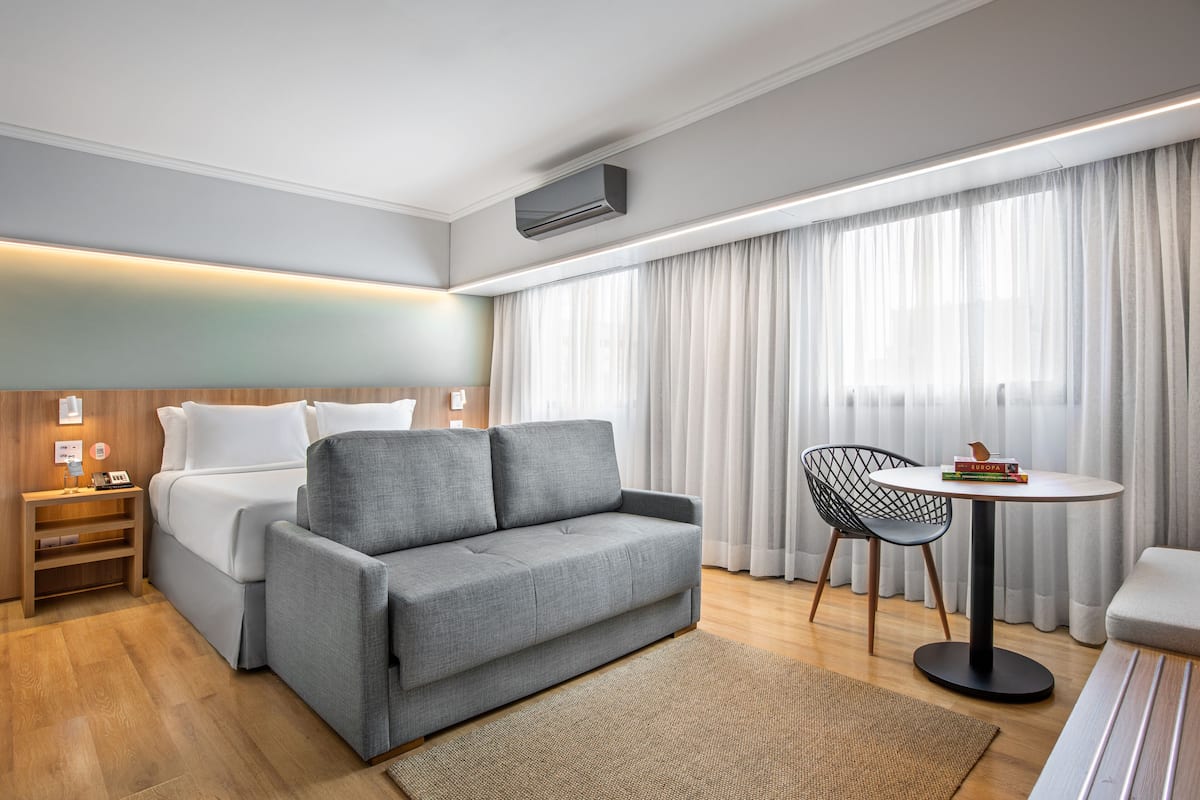 A modern hotel room with a grey sofa, a small round table, a chair, a bed, and a large window with curtains.