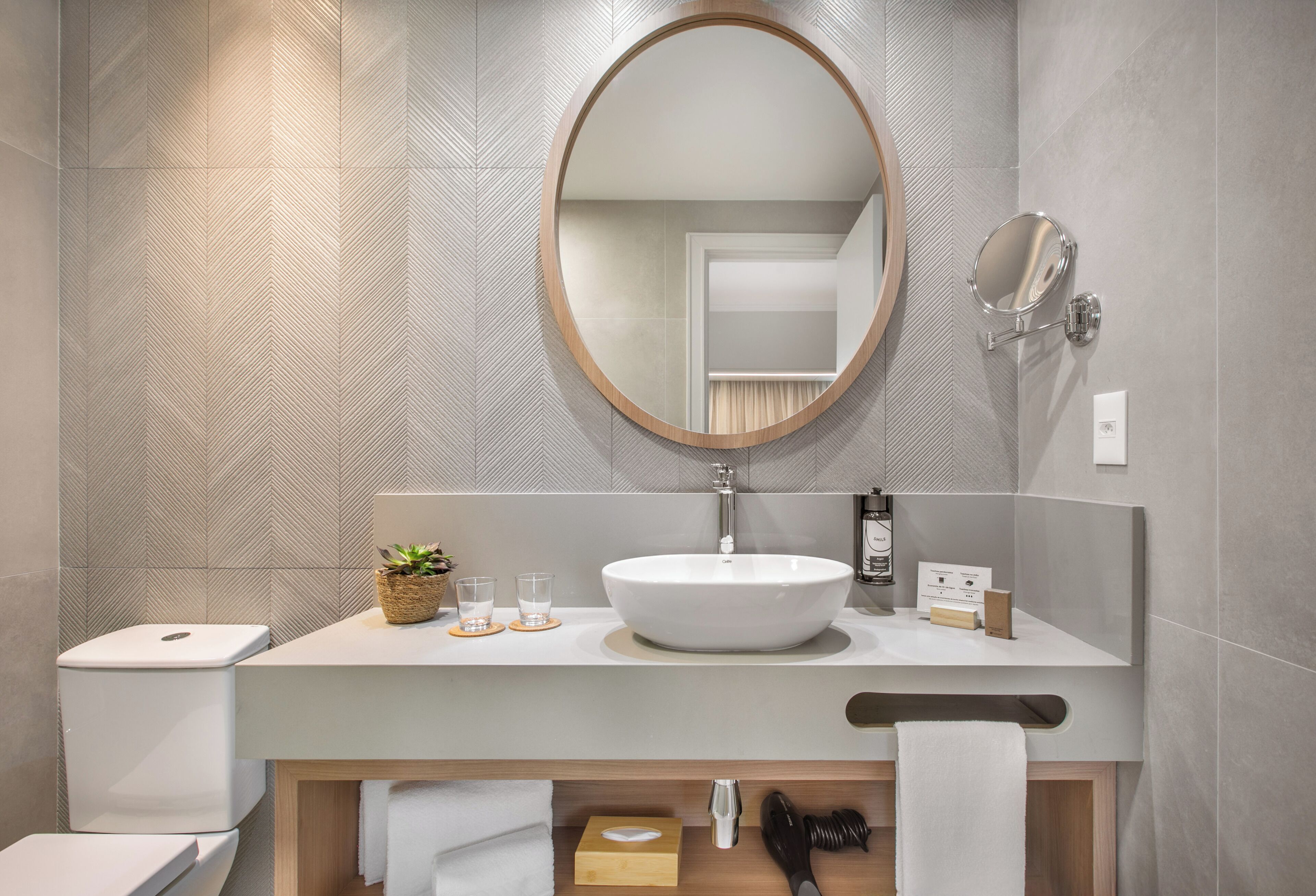Een moderne badkamer met een grote, ovale spiegel, een witte wastafel en een houten badkamerkast.