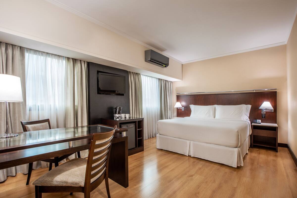 A hotel room with a bed, a desk with a chair, a television, and a nightstand with a lamp.