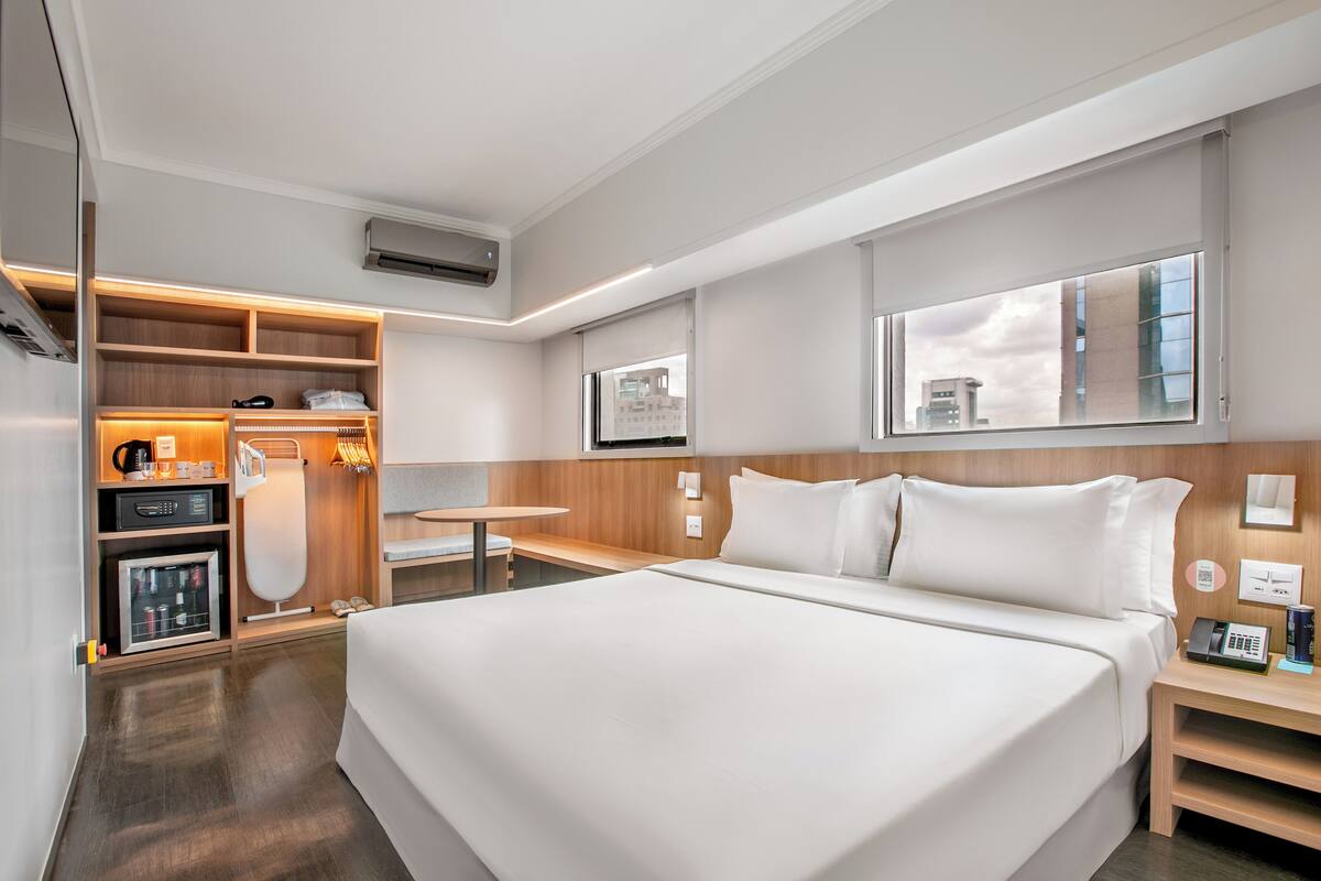A modern hotel room with a large bed, a built-in desk, a wardrobe, and a view of the city through a large window.