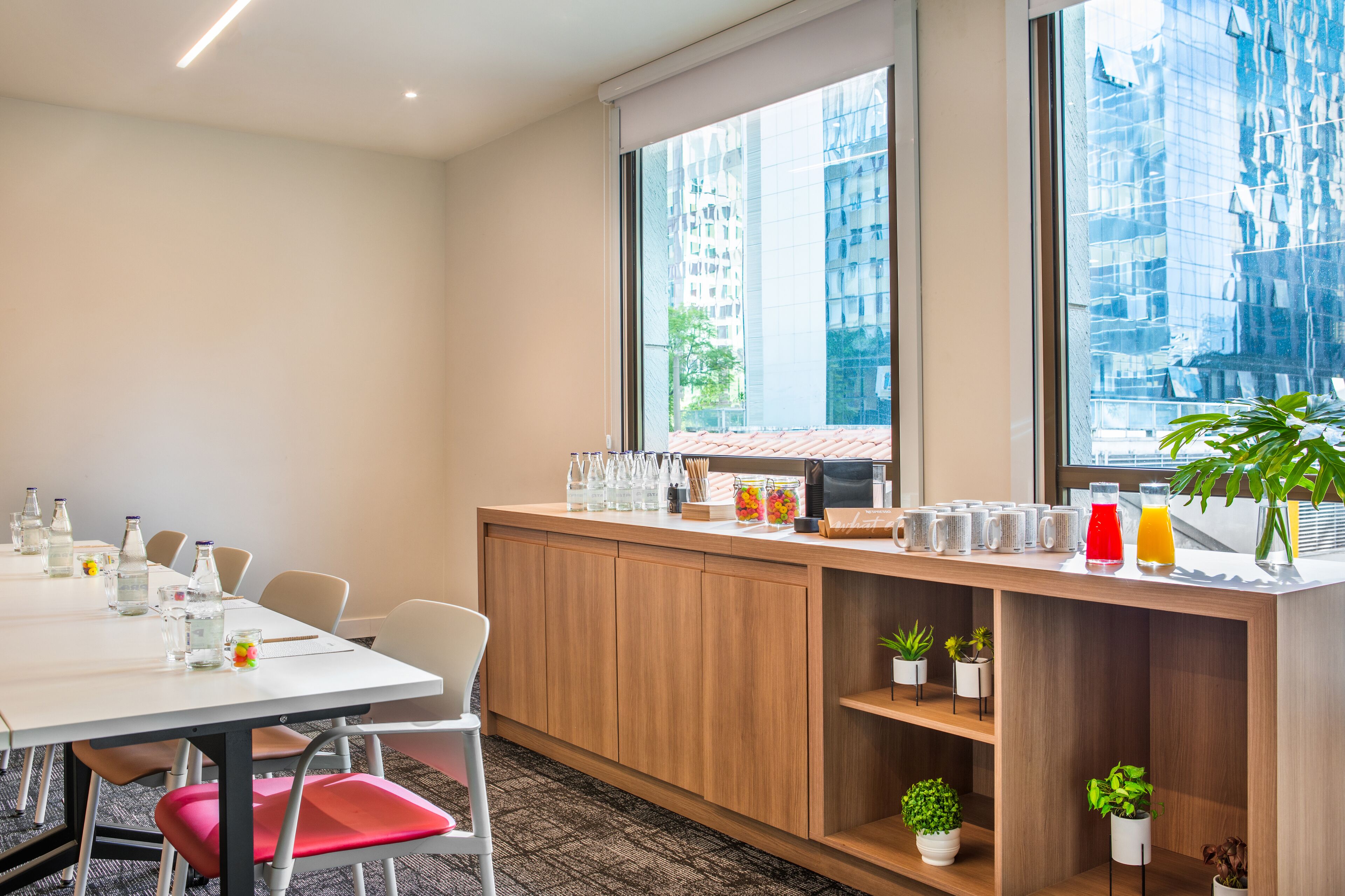 A conference room with a long table, chairs, a wooden counter with amenities, and large windows with a city view.