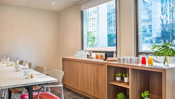 A conference room with a long table, chairs, a wooden counter with amenities, and large windows with a city view.