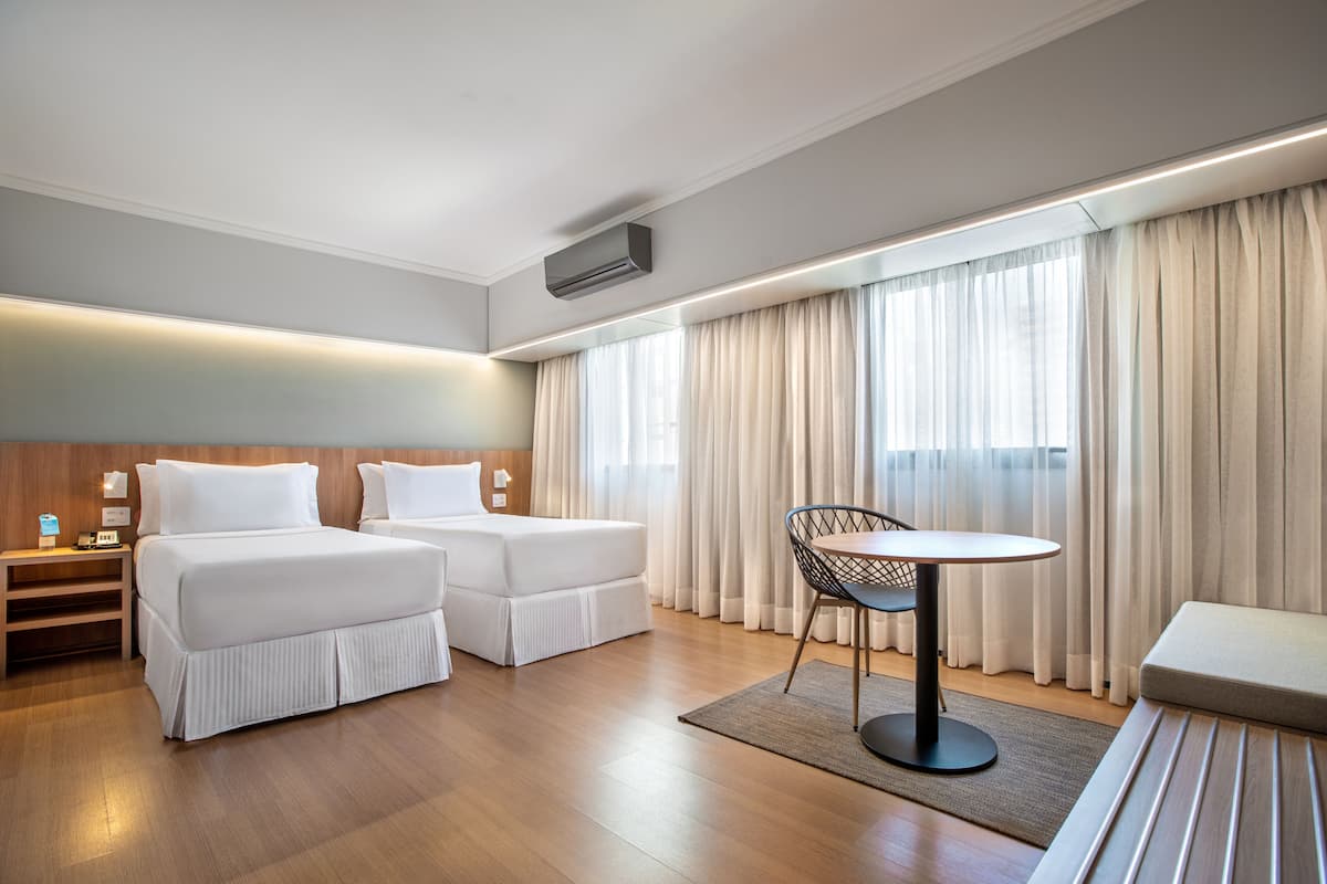 A hotel room with two beds, a small table, and a chair. There is a desk with a lamp and a phone. The room has wooden flooring and large windows with curtains.