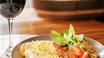 Lautasella on pastaa, paneroitua pihviä ja tomaattia. Annoksen kanssa tarjoillaan lasillinen punaviiniä. Ruokailutila on moderni, ja siinä on puinen pöytä sekä pehmustettu istuinalue taustalla.