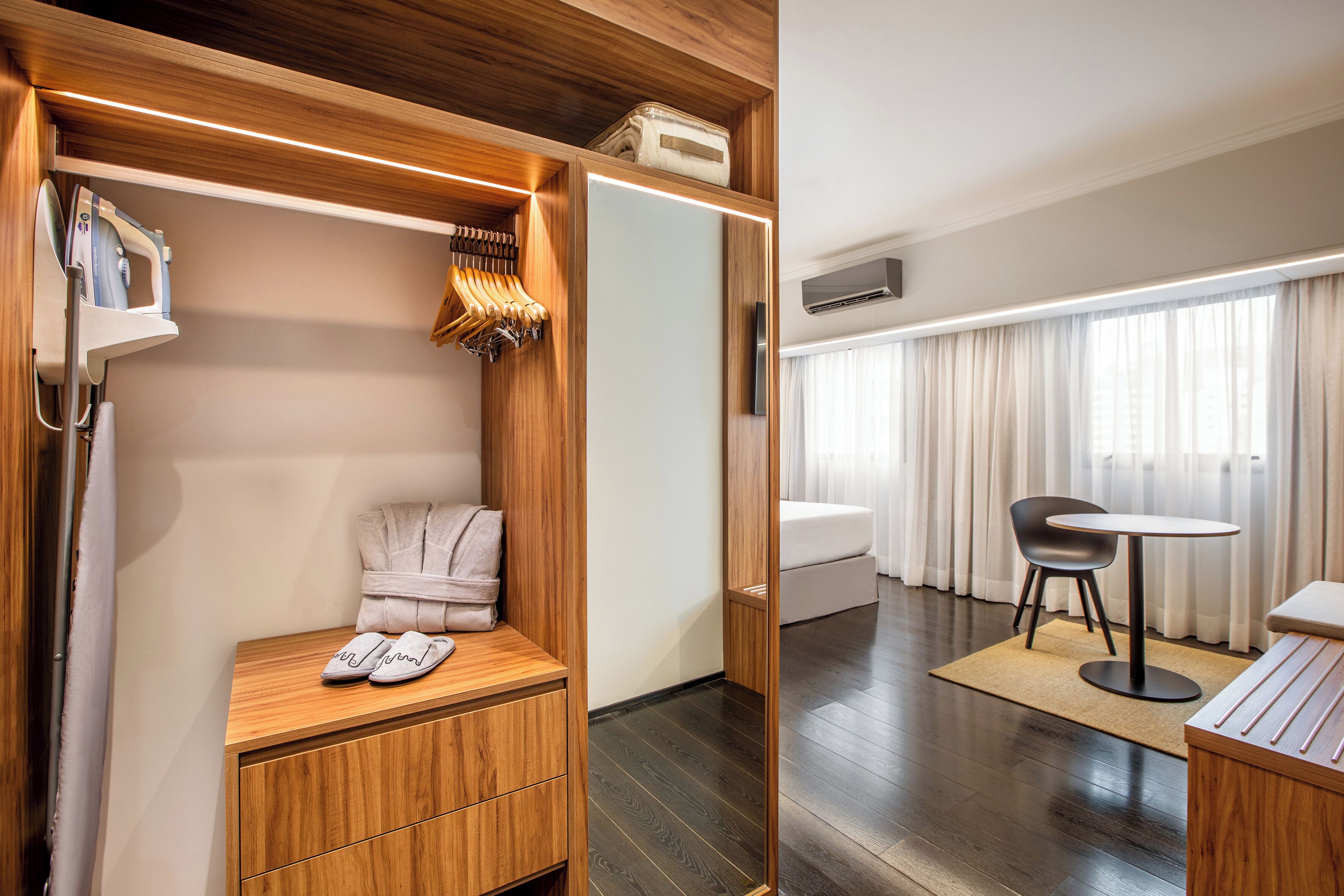 Habitación de hotel con tocador de madera, bata, pantuflas, una mesita, una silla y una cama.