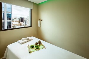 A hotel room with a large window, a bed with a white sheet, a towel, a small green tray with a bowl and a plant, and a wall-mounted towel rack.
