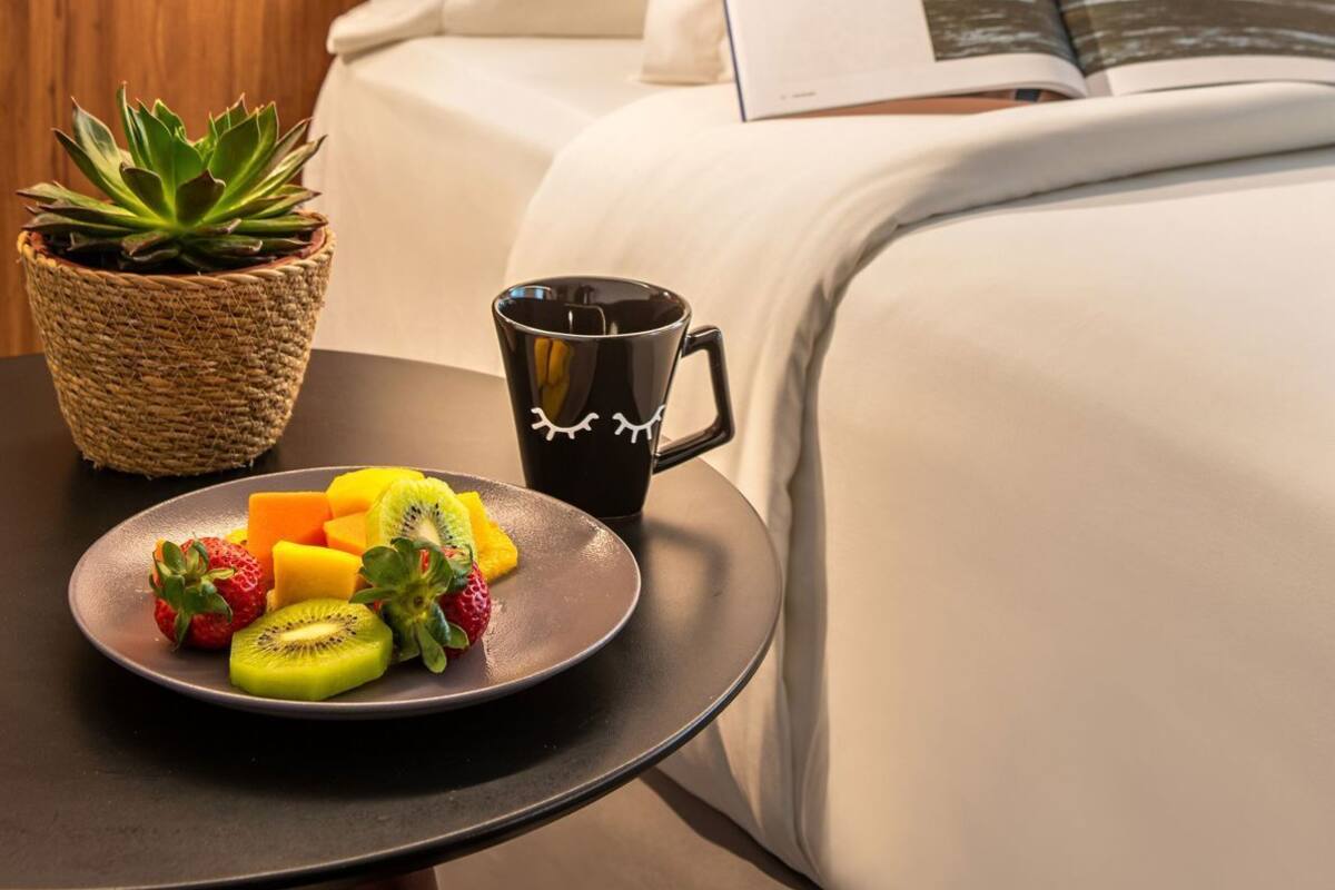 A neatly made bed with white linens, a book with a landscape image, a black mug, a plate of fruit, and a succulent plant on a bedside table.
