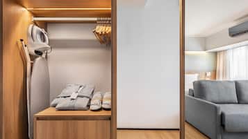 Ein moderner Kleiderschrank mit Kleiderstange, eine Holzkommode mit gefalteten Handtüchern und Hausschuhen sowie ein graues Sofa in einem Wohnbereich mit Fenster und Klimaanlage.