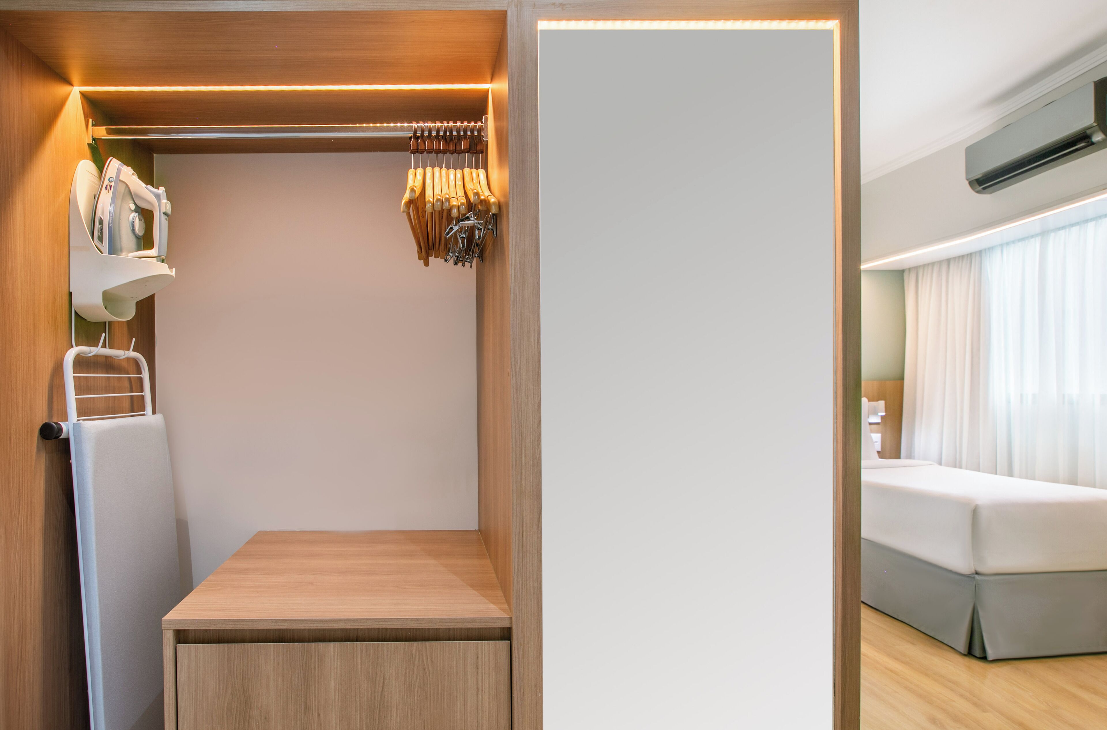 A compact room with a wooden bench, a built-in wardrobe with hanging clothes, and a bed with a grey skirt.