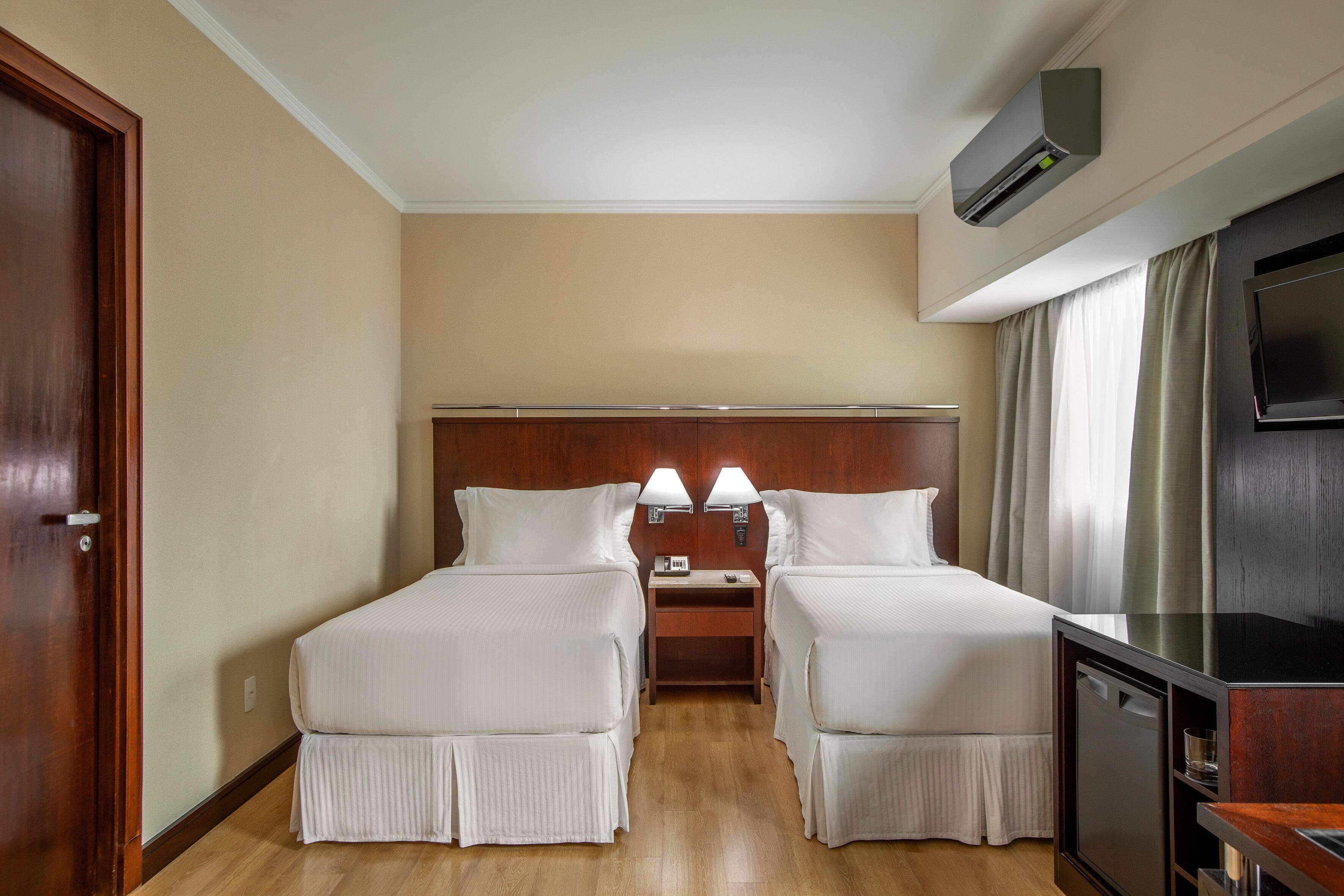 A hotel room with two beds, a nightstand, a TV, and an air conditioning unit.
