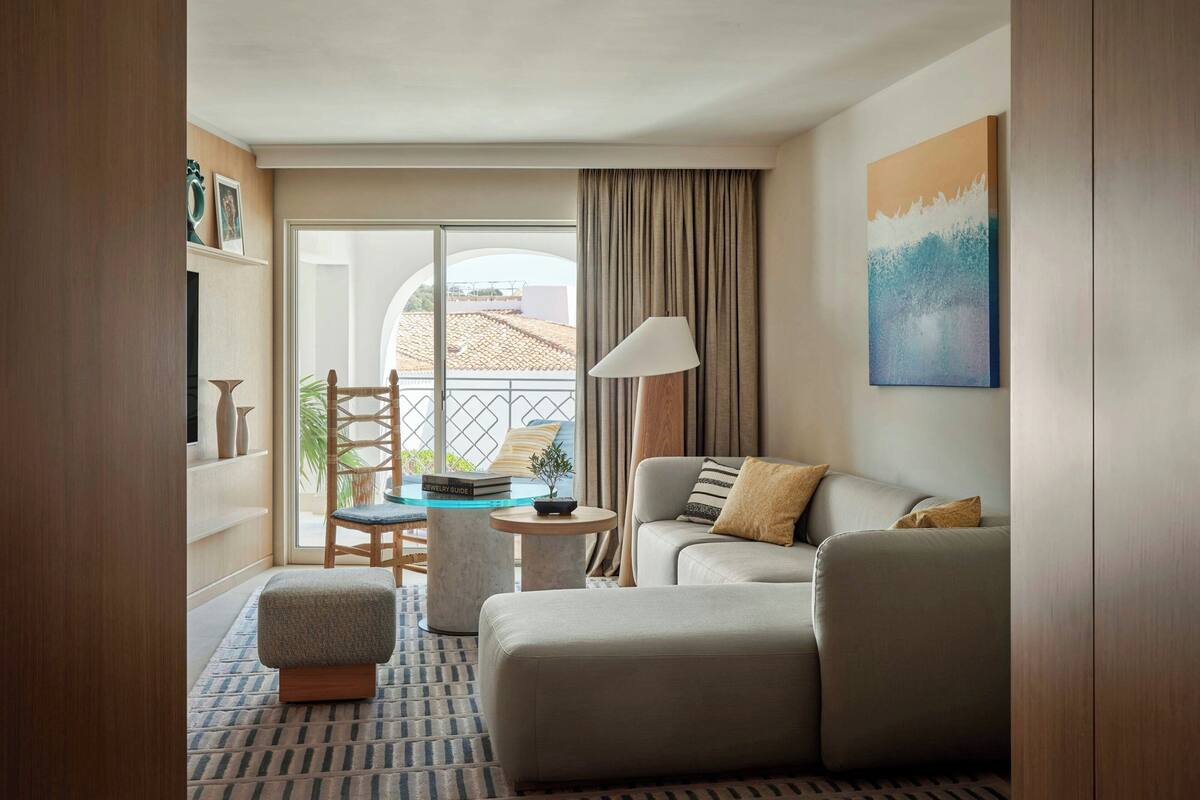 A modern hotel room with a sofa, ottoman, and a small table with a lamp. There is a balcony with a view of buildings.
