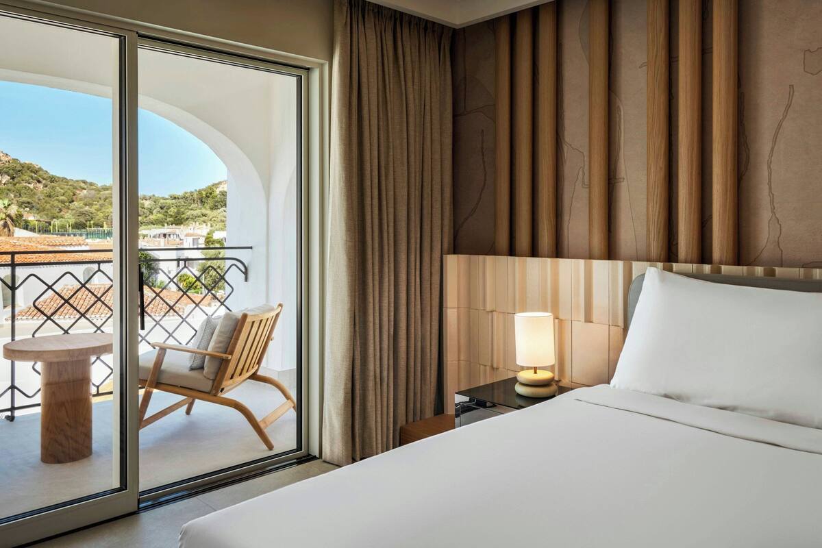 A hotel room with a large bed, a nightstand with a lamp, a chair by the window, and a view of the outdoors.