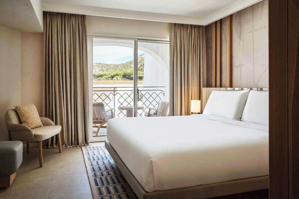 A hotel room with a large bed, a chair, a small table, and a balcony with a view of the landscape.