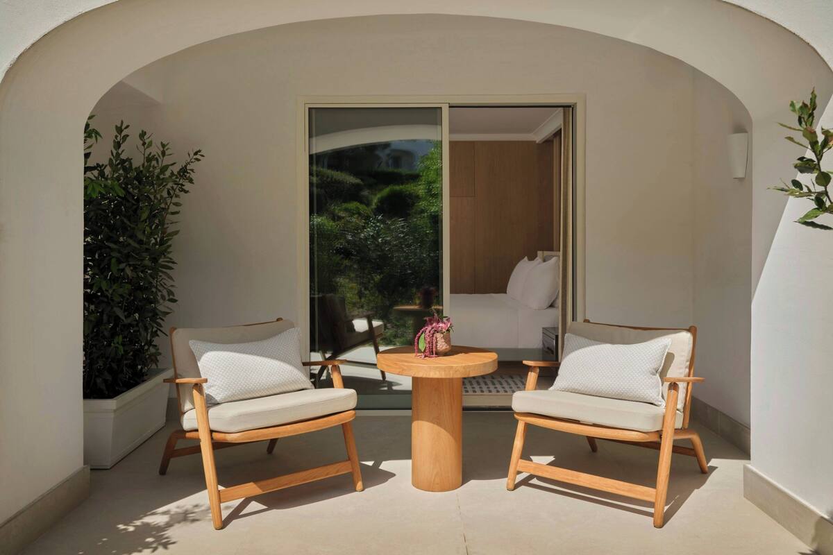A balcony with two wooden chairs, a round wooden table, and a glass door leading to a room.