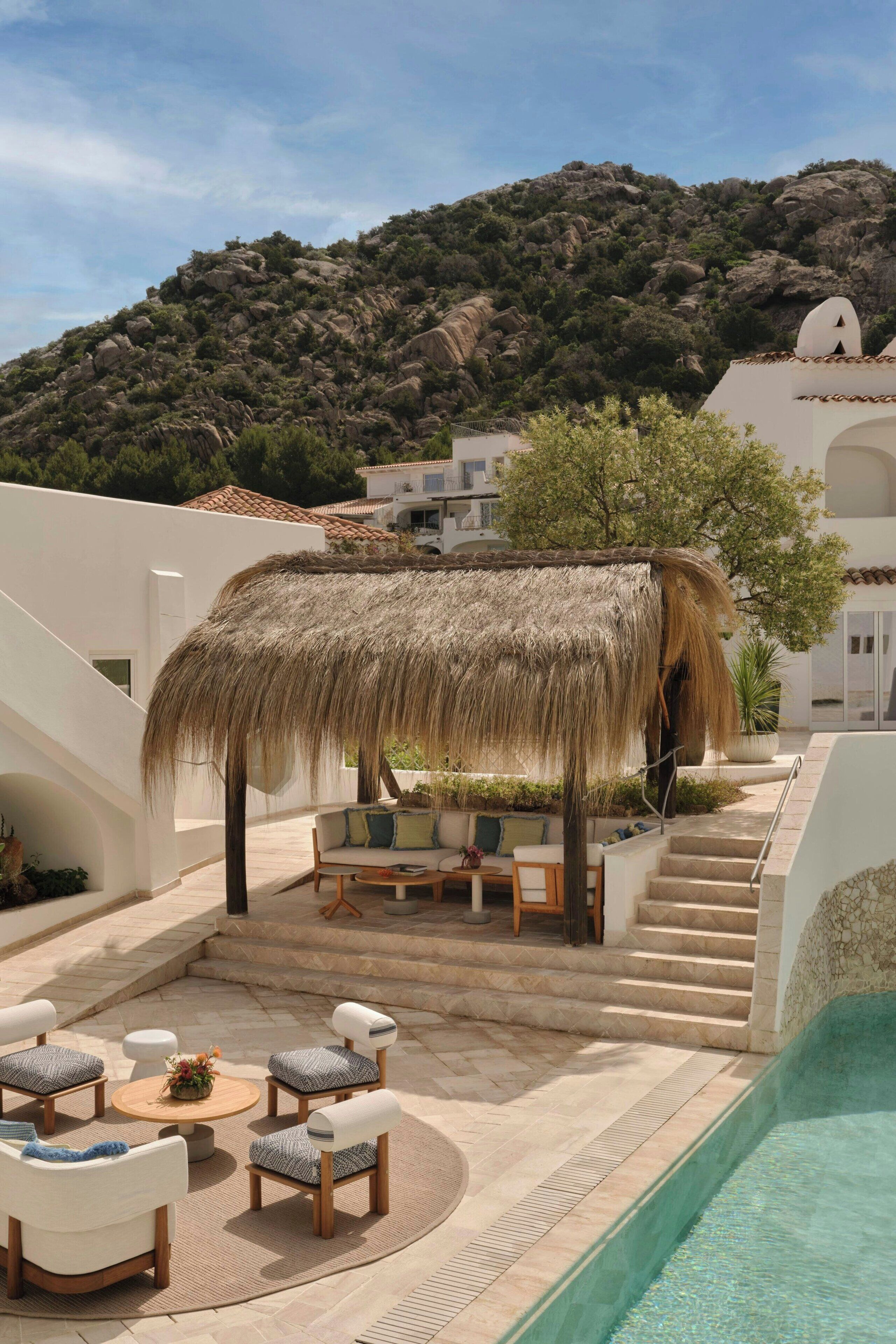A poolside area with outdoor seating, a thatched roof structure, and a mountainous backdrop.