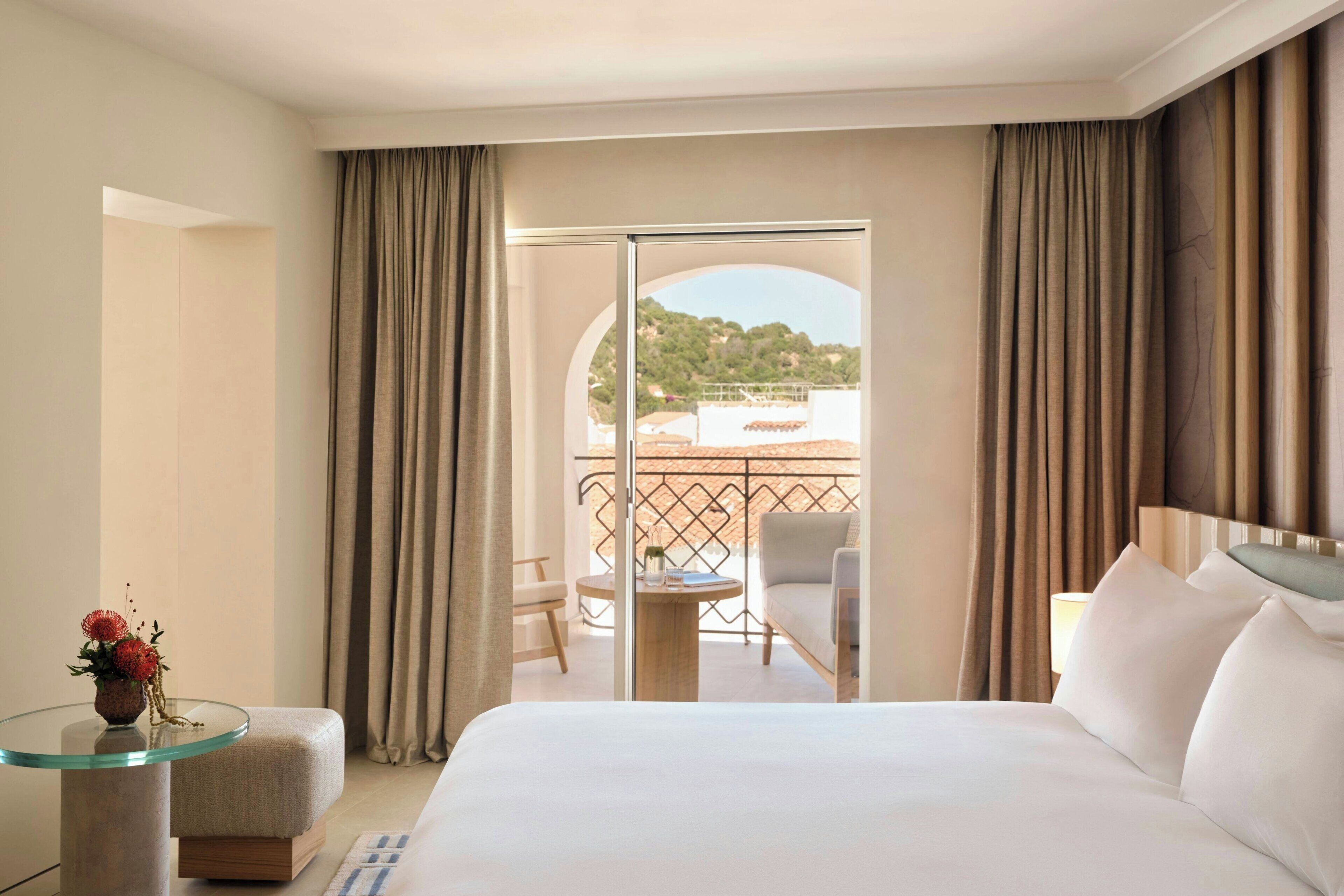 A hotel room with a bed, a glass table with a vase, and a view of a balcony with chairs and a table.