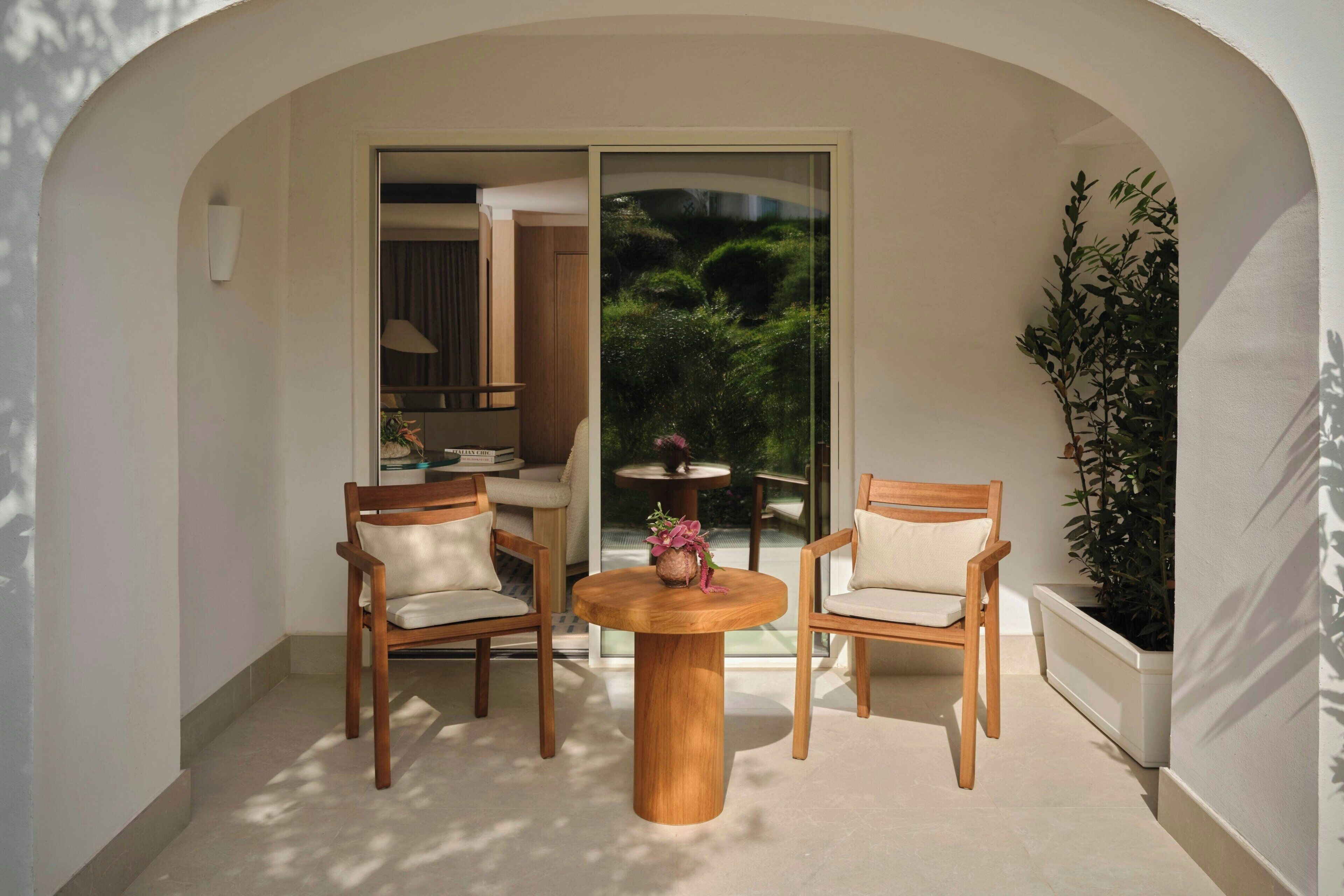 A balcony with a round wooden table and two chairs, a potted plant, and a view of a garden through a glass door.